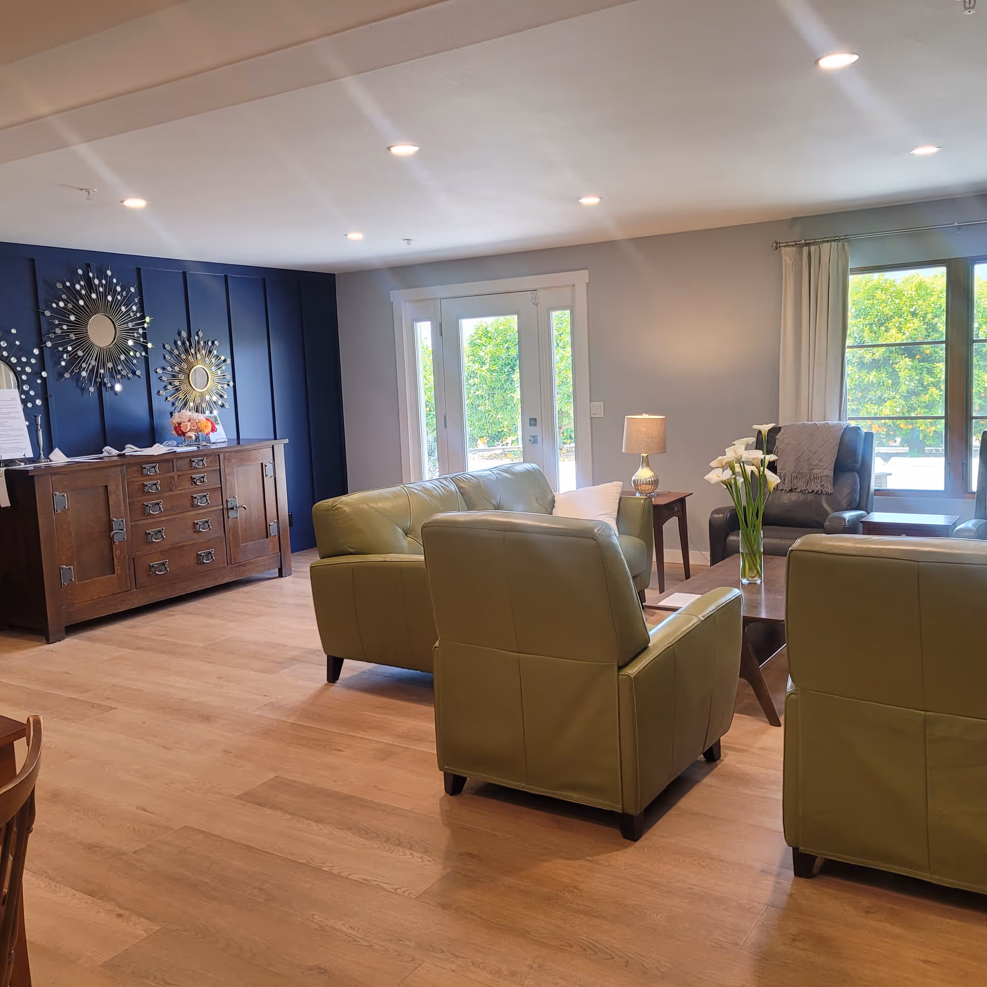 A bright communal living room with green leather armchairs around a coffee table, a wooden sideboard with decorative mirrors, and windows/doors letting in natural light.