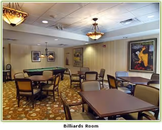 A billiards room with a pool table in the center surrounded by several tables and chairs. The room has patterned carpet, beige walls with framed artwork, and ceiling lights including two hanging chandeliers.