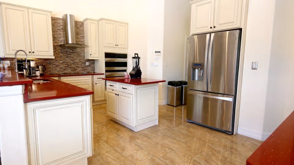 A modern kitchen with cream-colored cabinets and red countertops. The kitchen features a stainless steel refrigerator with a water dispenser, a built-in oven, a blender on a small island, a coffee maker, and a stainless steel range hood above the stove. The floor is tiled with a glossy finish, and there are two trash bins against the wall.