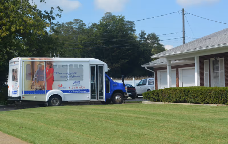 A small white and blue shuttle bus parked on a grassy area next to a brick building with white trim and a covered porch. The bus has an image of a caregiver assisting an elderly person and text that reads 'Where caring people make the difference! Mount Vernon 812-838-6554 ASCseniorCare.com'. Trees and a clear sky are visible in the background.