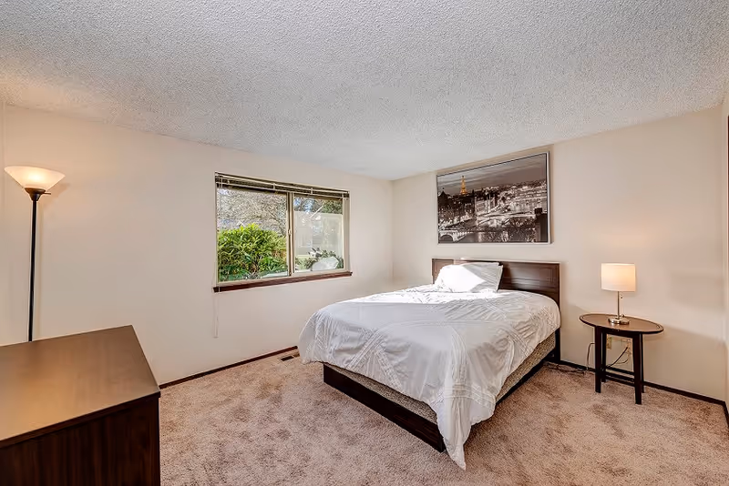 A bedroom with a bed covered in white bedding, a wooden headboard, a round side table with a lamp, a wooden dresser, a floor lamp, a window showing greenery outside, and a black and white framed picture on the wall.