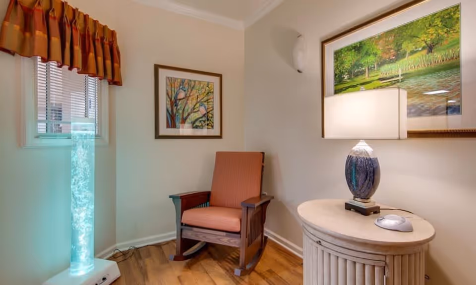 A cozy corner of a room featuring a wooden rocking chair with orange cushions, a small round side table with a decorative lamp, a tall illuminated bubble lamp near a window with blinds and an orange valance, and two framed paintings on the walls.