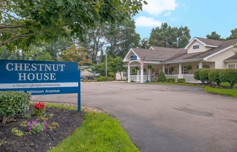 Exterior view of Chestnut House, a senior lifestyle community, showing a single-story building with a covered entrance, an American flag, and surrounding greenery. A blue sign in the foreground reads 'Chestnut House, a Senior Lifestyle community, Johnson Avenue.'