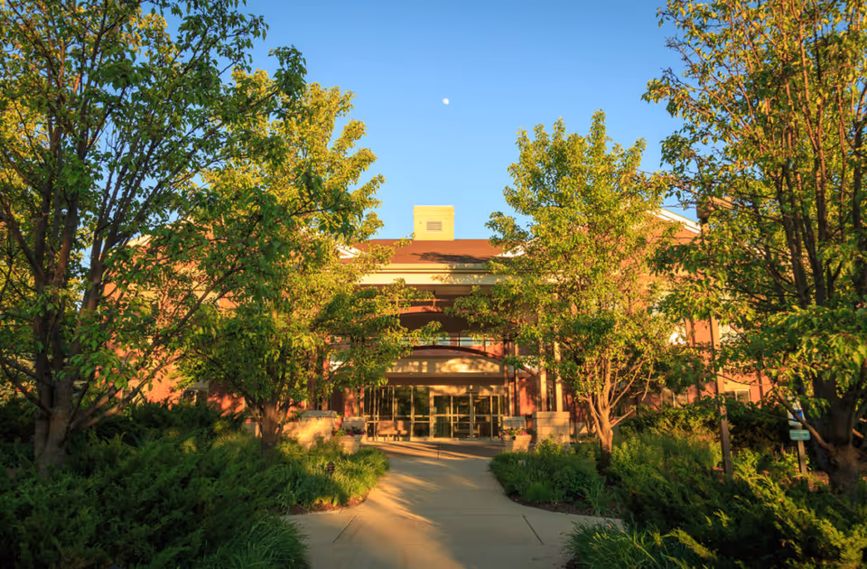 Entrance to a senior living facility named Sedgebrook, framed by lush green trees and shrubs on either side of a paved walkway under a clear blue sky with the moon visible.