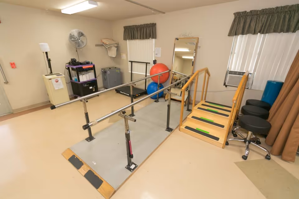 A physical therapy room with parallel bars for walking exercises, a small wooden staircase with handrails, exercise balls, a large mirror, stools, and various equipment against the walls.