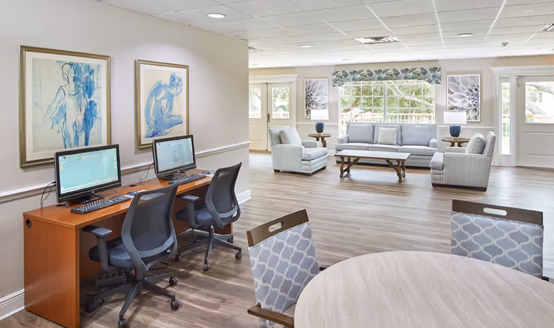 Bright communal lounge with a computer workstation, round table, and a seating area of sofas and armchairs.
