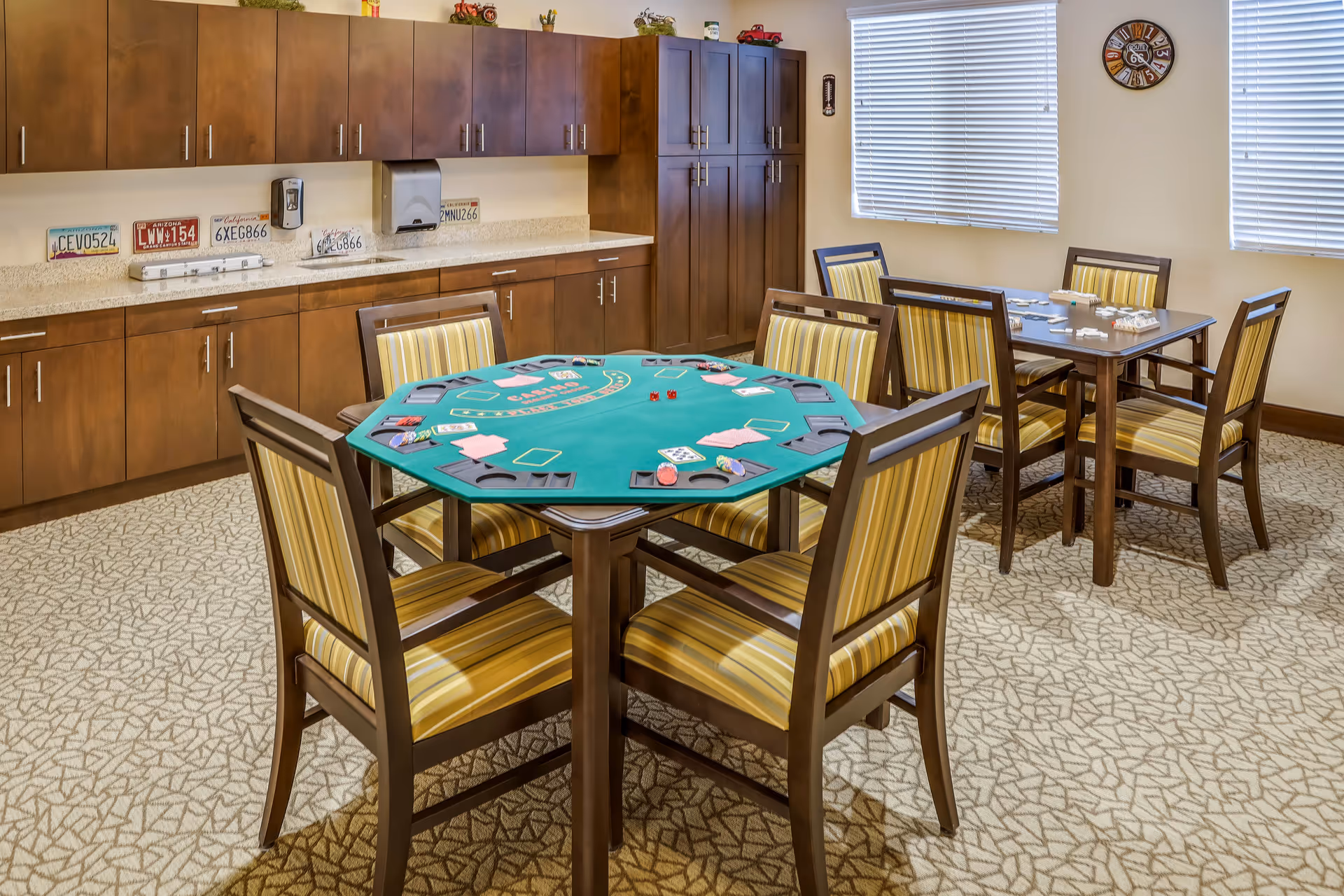 A well-lit game room with two tables and several chairs. The table in the foreground is set up for a card game with poker chips, cards, and dice on a green felt surface. The table in the background has a game of dominoes in progress. The room features wooden cabinets along one wall, decorative license plates, and a clock on the wall between two windows with blinds.