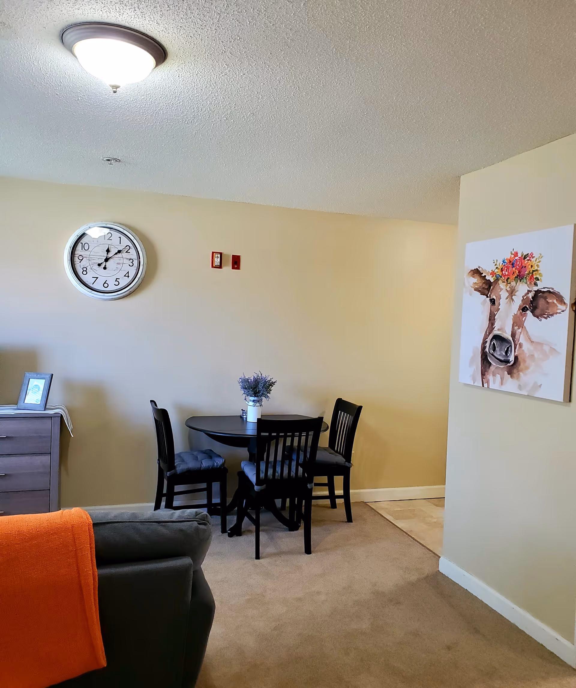 A small dining area with a round black table and three black chairs with cushions. A vase with purple flowers is on the table. On the wall above the table is a round clock showing the time as 12:15. To the right, there is a painting of a cow with a flower crown. Part of a gray couch with an orange blanket draped over the back is visible in the foreground. The walls are beige and the floor is carpeted.