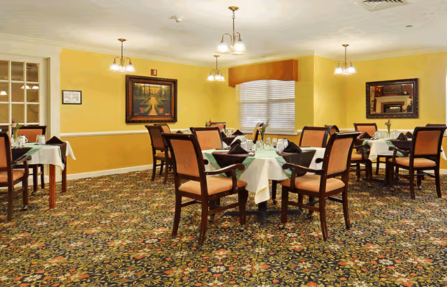 A dining room with several tables covered with white tablecloths and set with glasses, napkins, and silverware. The room has yellow walls, a patterned carpet, framed artwork on the walls, and multiple ceiling light fixtures. There is a window with blinds and a valance.