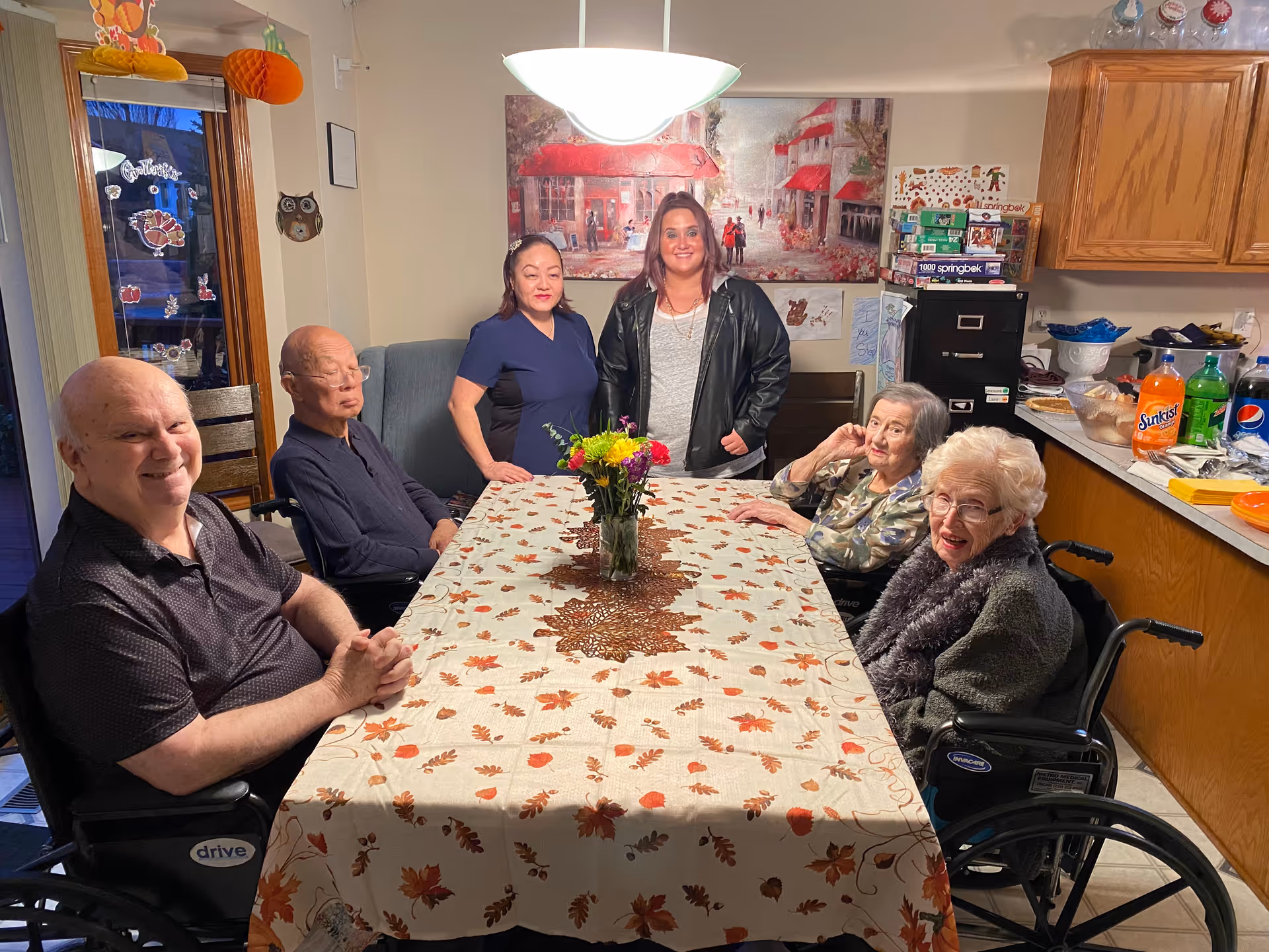 A group of five elderly people and two caregivers gathered around a dining table covered with a fall-themed tablecloth. The table has a vase with colorful flowers in the center. The room has wooden cabinets, a countertop with snacks and drinks, and a painting on the wall. Some of the elderly individuals are seated in wheelchairs.