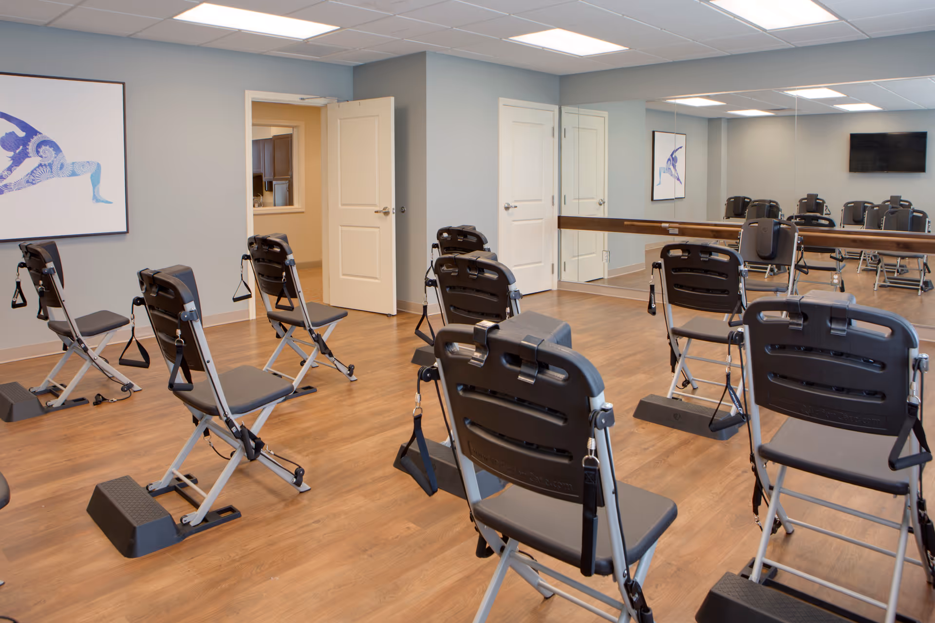 Exercise room with several black folding chairs equipped with resistance bands arranged on a wooden floor. The room has light gray walls, a large mirror on one side, a wall-mounted TV, and a framed artwork of a person in a yoga pose. An open door leads to another room.