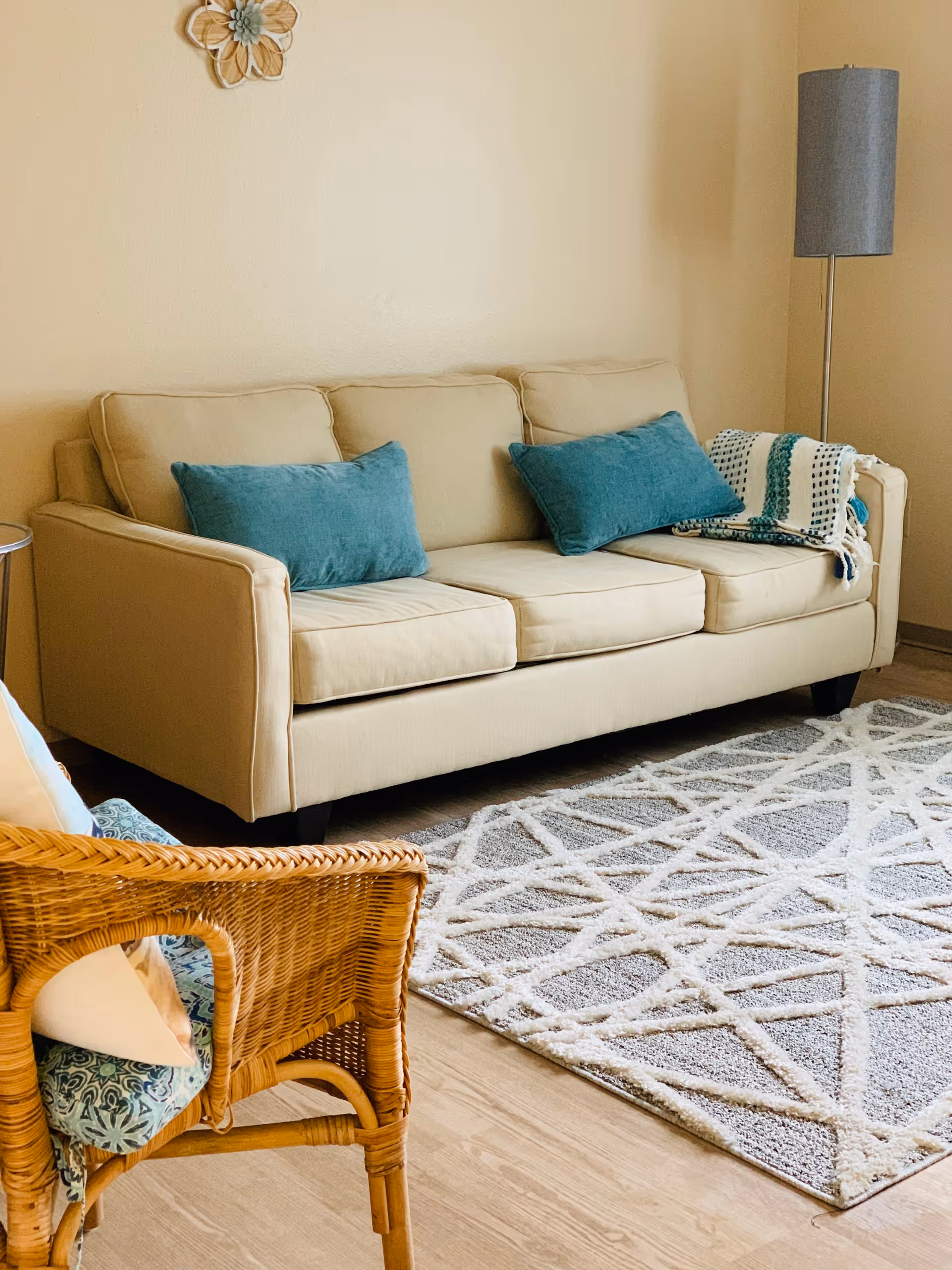 A cozy living room area featuring a beige sofa with two teal cushions and a teal and white throw blanket on the right armrest. In front of the sofa is a light gray rug with a white geometric pattern. To the left is a wicker chair with a blue patterned cushion and a white pillow. A tall gray floor lamp stands in the corner against a beige wall, which is decorated with a small floral wall ornament.