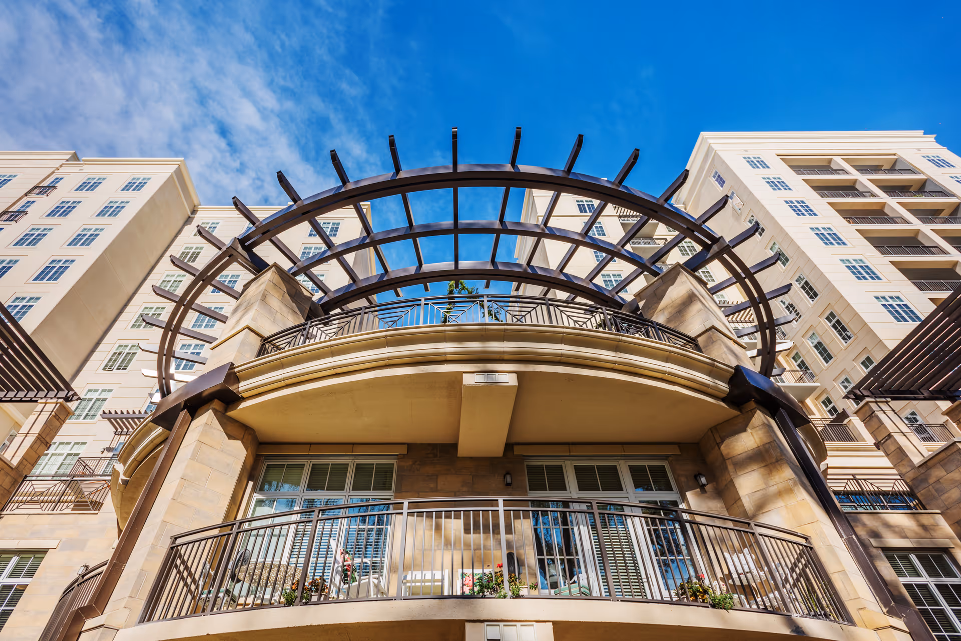 Low-angle view of a multi-story building facade with balconies and a decorative metal pergola against a blue sky.