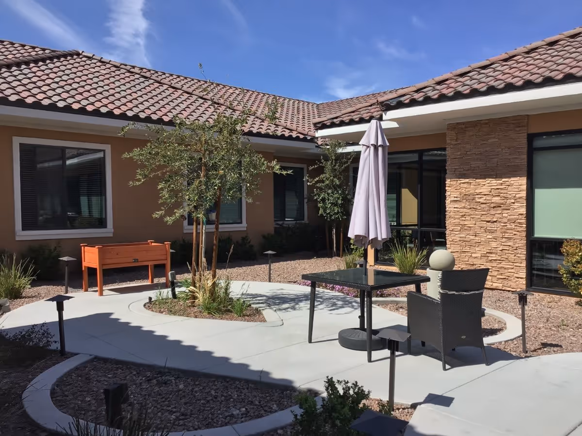 Outdoor courtyard area at Grand Montecito Memory Care with a tiled roof building surrounding it. The courtyard features a small tree, a glass-top table with a closed umbrella, a black chair, and a raised wooden planter box. The ground is covered with concrete walkways and gravel, with some small plants and shrubs around.