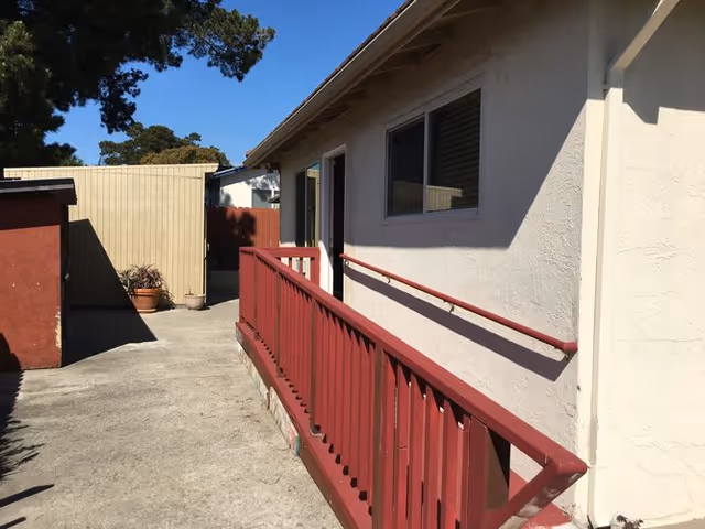 Outdoor walkway alongside a beige building with a red wooden handrail and railing. There are potted plants near a beige fence at the end of the walkway, and trees are visible in the background under a clear blue sky.