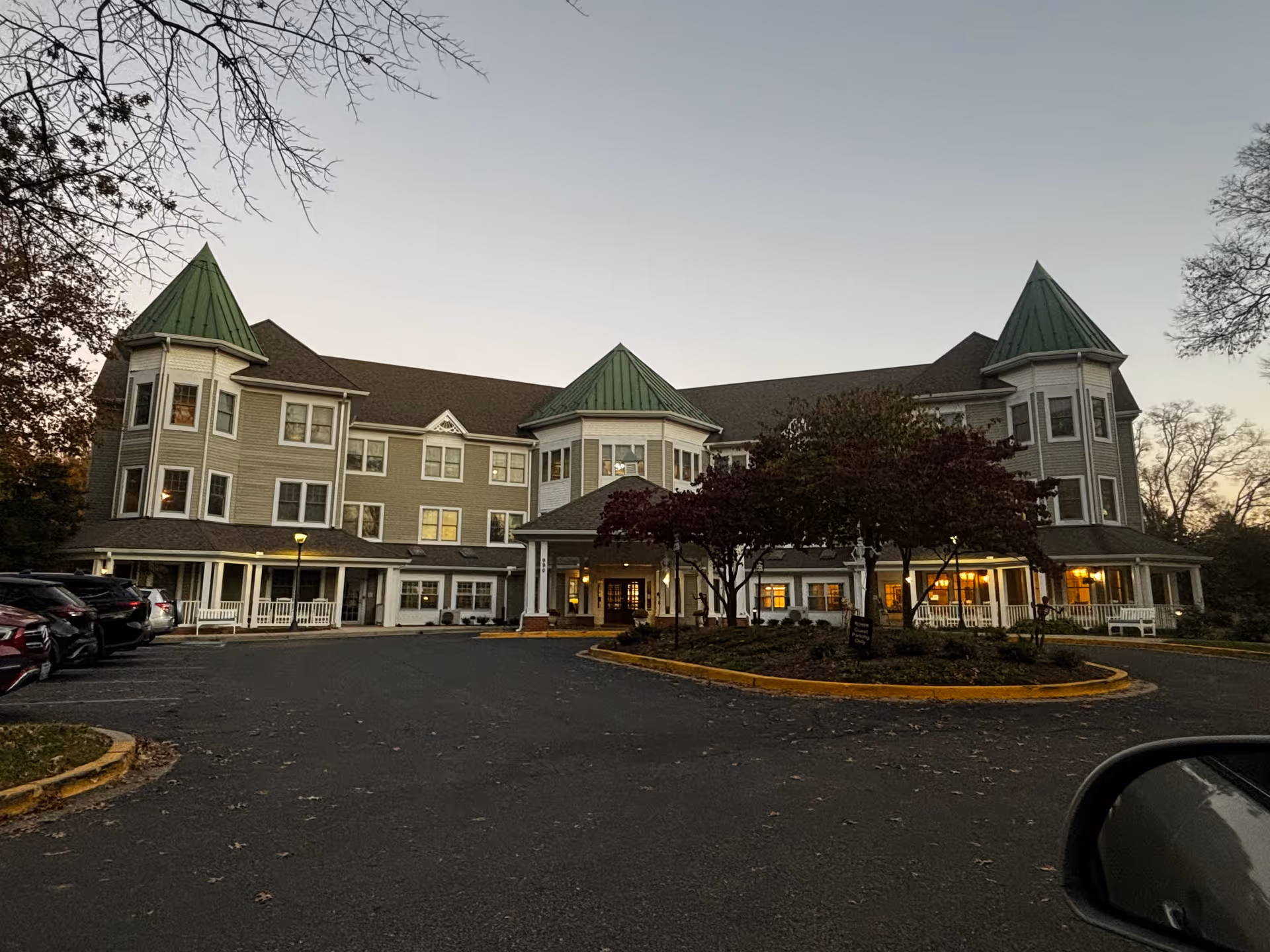 Three-story senior living building with green turret roofs, lit windows, and a circular driveway at dusk.