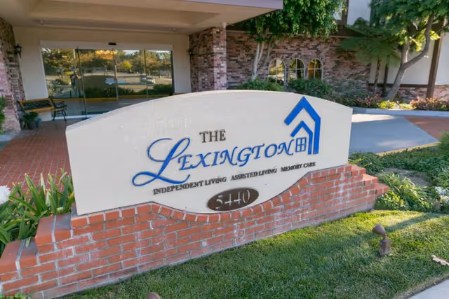 Outdoor view of the entrance sign for The Lexington assisted living facility, featuring a brick base and white curved sign with blue and black text, surrounded by greenery and part of the building visible in the background.