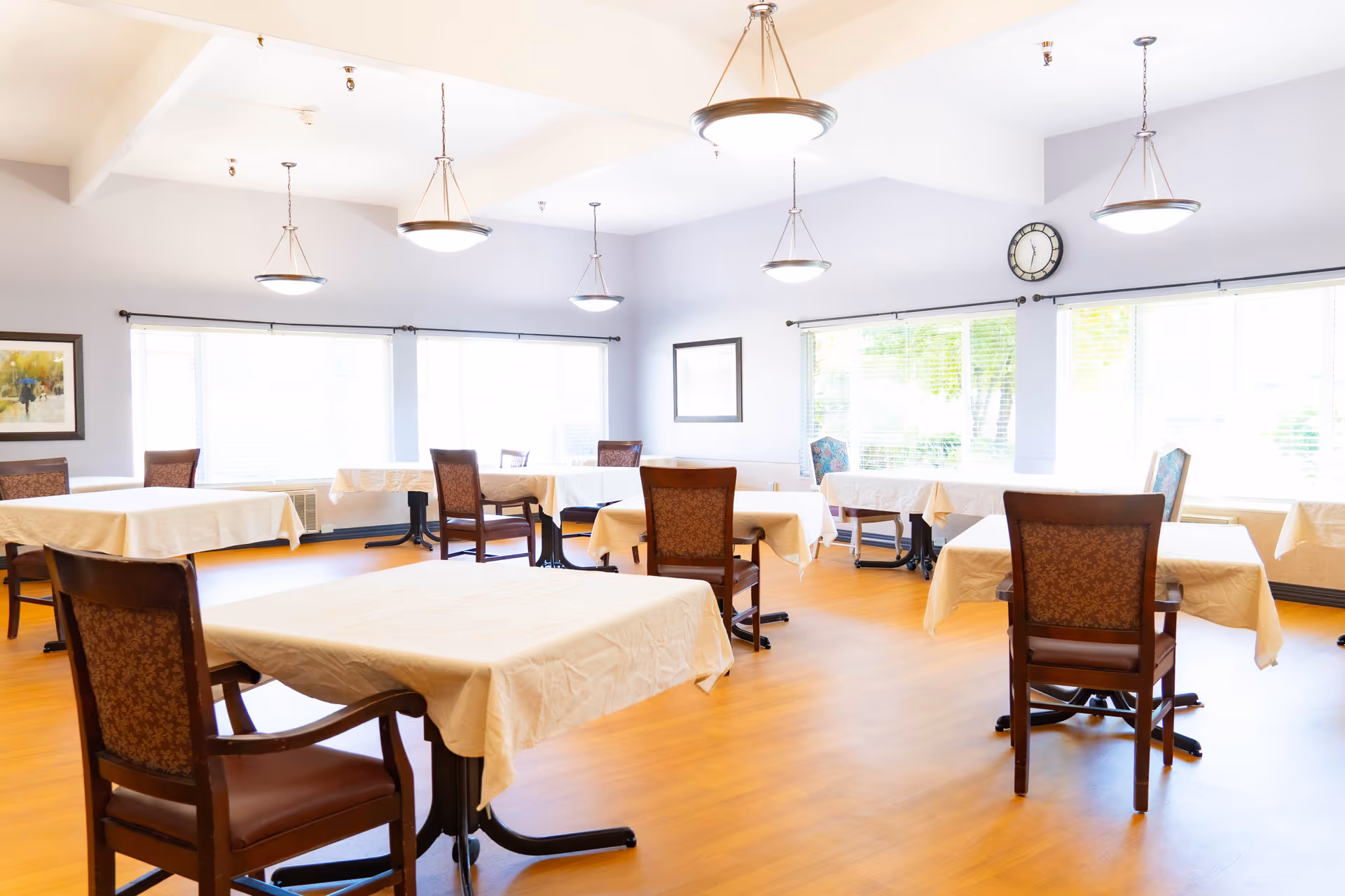 Bright dining room with multiple tables covered in white tablecloths and wooden chairs arranged under pendant lights and large windows.