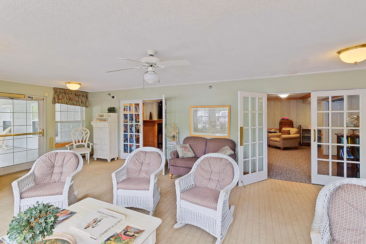 A bright and cozy living room in a senior housing facility featuring four white wicker armchairs with pink cushions arranged around a white coffee table with books and a small plant. There is a purple loveseat against the wall with two floral pillows, a white cabinet, and a white chair near a window with floral valance. French doors lead to another room with additional seating and a globe on a stand. The room has beige carpeting, light green walls, and a ceiling fan with a light fixture.