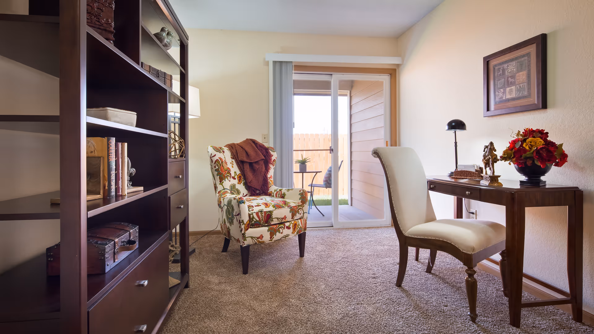 A cozy interior room with a floral upholstered armchair draped with a brown throw, a wooden desk with a beige cushioned chair, a table lamp, decorative items, and a framed picture on the wall. There is a sliding glass door leading to a small outdoor patio with a table and chairs. A dark wooden bookshelf with books and decorative boxes is on the left side of the room.