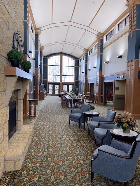 A spacious and well-lit common area with a high vaulted ceiling, large windows at the far end, and a patterned carpet. The room features a stone fireplace on the left with potted plants on the mantel, several blue upholstered armchairs with small round tables on the right, and a long wooden table with chairs in the center. The walls have wood paneling and sconces providing light.