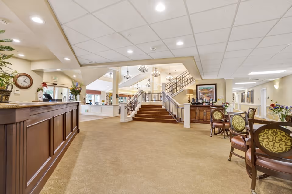 Spacious and well-lit interior of a senior living facility lobby with a wooden reception desk on the left, carpeted floor, a staircase with metal railings leading to an upper level, several chandeliers hanging from the ceiling, and a seating area with round tables and chairs on the right. There are decorative plants, framed artwork, and a clock on the wall near the entrance.