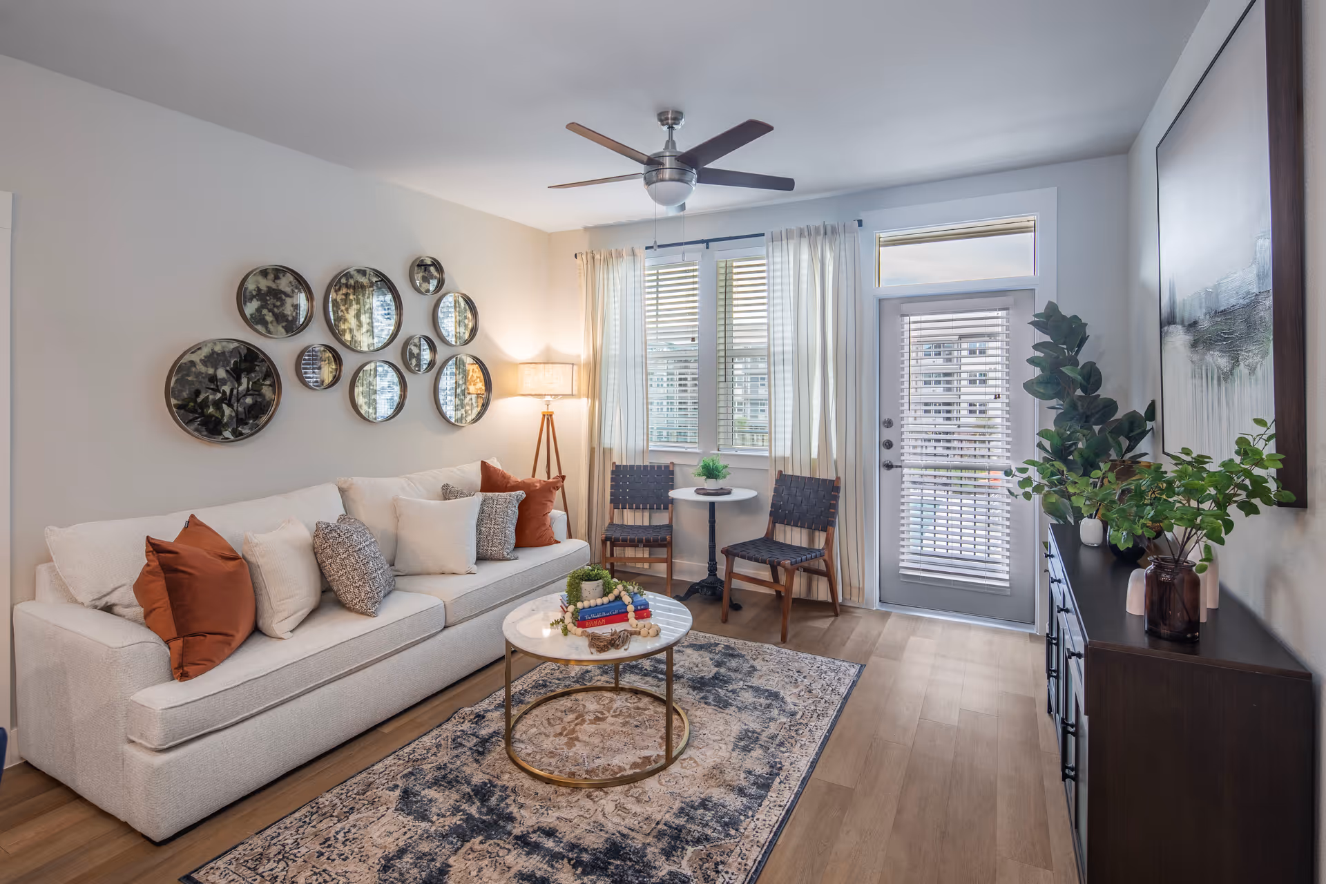 A modern living room with a white sofa adorned with multiple cushions in neutral and rust colors. A round coffee table with decorative items sits on a patterned rug. On the wall above the sofa, there are multiple round mirrors of varying sizes arranged in a cluster. Two woven chairs and a small round table are positioned near a window and a glass door with blinds. A dark wooden cabinet with plants and decorative items is against the opposite wall, which also features a large framed artwork. The room has wooden flooring, a ceiling fan with lights, and sheer curtains on the windows.