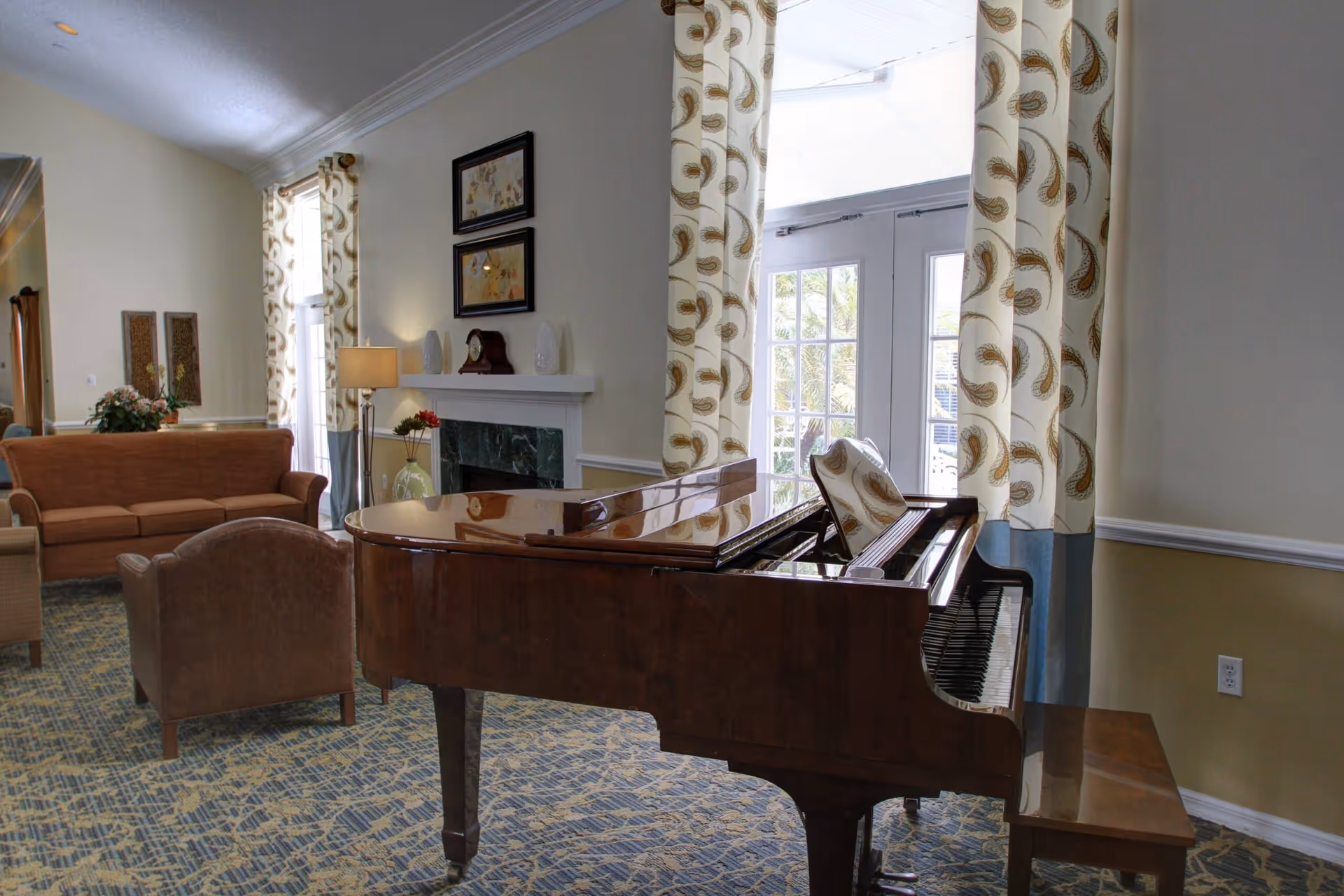 A bright and cozy living room area in a senior living facility featuring a polished grand piano with a matching bench, a brown sofa, an armchair, a floor lamp, and a fireplace with decorative items on the mantel. Large windows with patterned curtains allow natural light to fill the room.