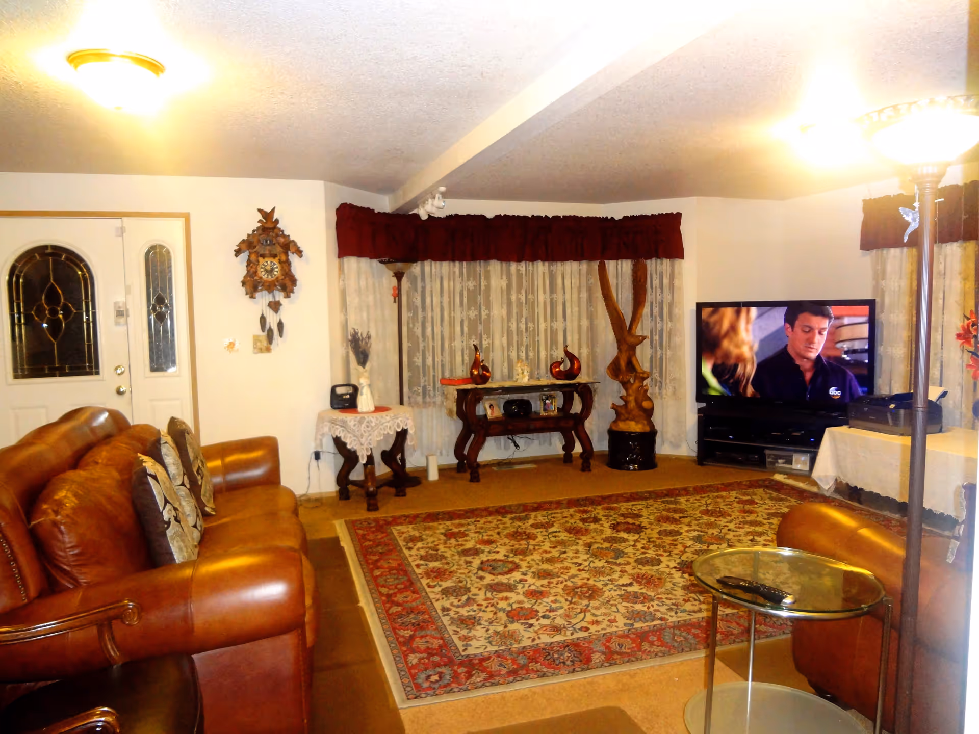 A cozy living room with a brown leather sofa, a patterned area rug, a glass side table with a remote control, a TV showing a scene from a show, decorative wooden sculptures, a cuckoo clock on the wall, and lace curtains with dark red valances.