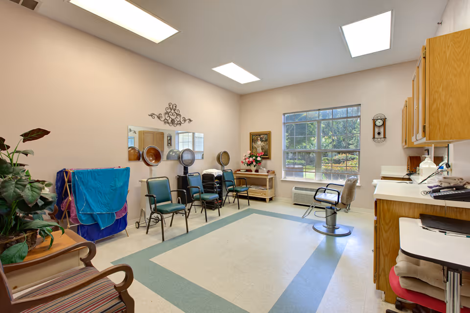 A bright salon room with three green chairs under hair dryers, a single salon chair near a large window, wooden cabinets on the right, a small table with flowers, a wall clock, and a decorative mirror on the wall. There is also a plant on a wooden chair and a drying rack with towels.