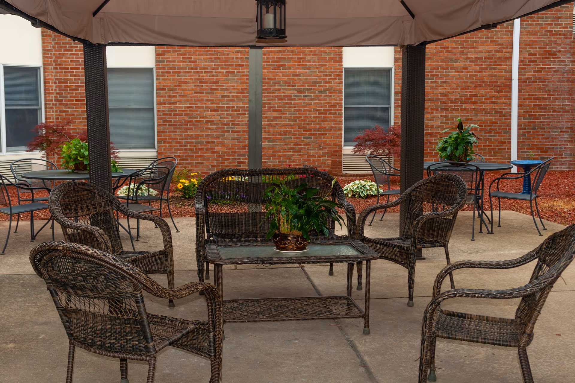 Outdoor seating area with a canopy overhead, featuring a set of dark wicker furniture including a loveseat, four chairs, and a glass-top coffee table with a potted plant. In the background, there are additional metal tables and chairs, some plants, and a brick building wall with windows.