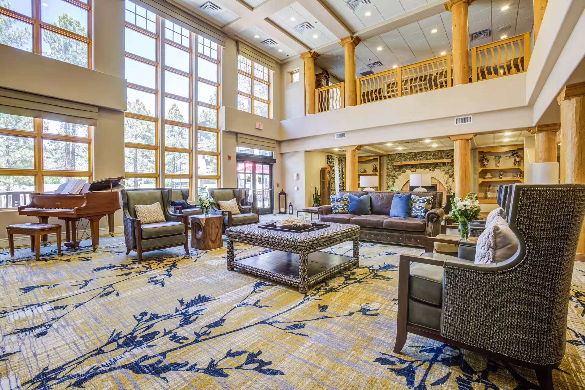 A spacious and well-lit senior living community lounge with large floor-to-ceiling windows, comfortable seating including armchairs and a sofa with cushions, a grand piano, wooden columns, and a patterned carpet. The room has a high ceiling with a balcony railing visible on the upper level and a stone fireplace in the background.