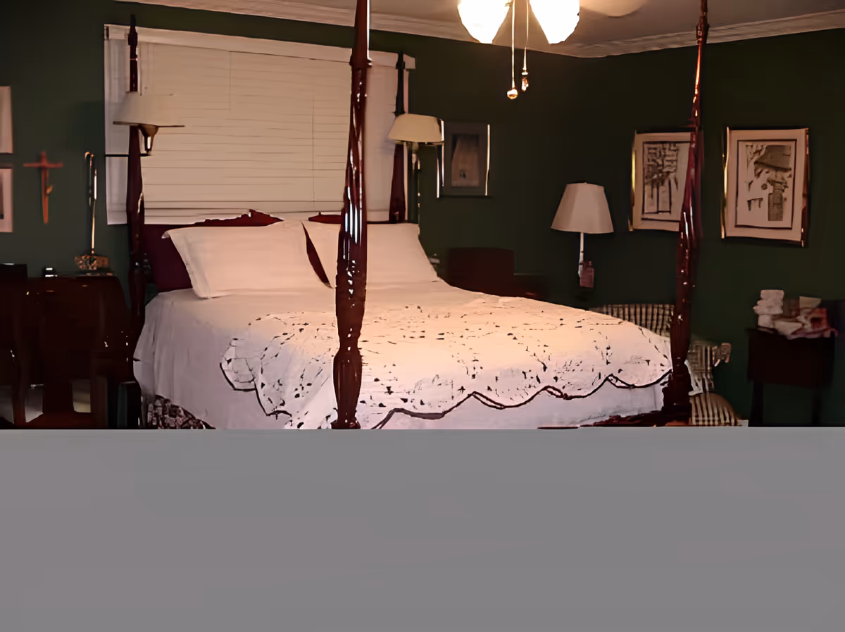 A bedroom with a four-poster bed covered with a white quilt. The walls are painted dark green and decorated with framed artwork. There are bedside tables with lamps on each side of the bed, and a window with closed blinds behind the bed.