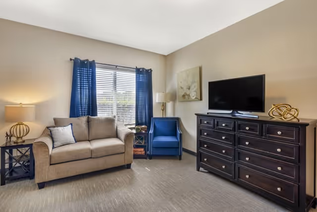 Well-lit living room with a loveseat, blue armchair, window with blue curtains, and a dark wood dresser topped by a TV.