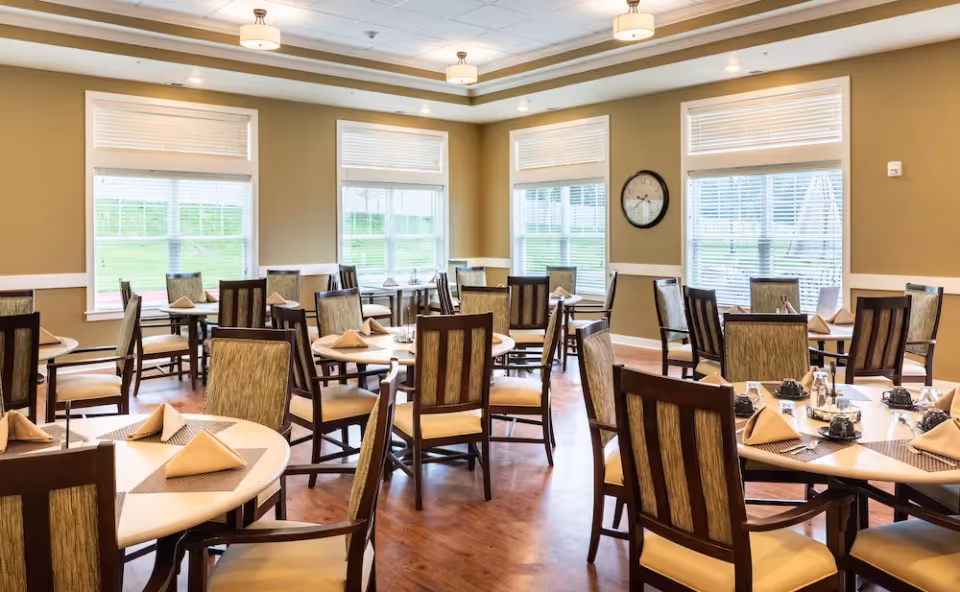 A bright dining room with multiple round tables set with beige napkins and placemats, surrounded by wooden chairs with cushioned seats and backs. Large windows with white blinds allow natural light to fill the room, and a clock is mounted on one of the beige walls. The floor is wooden, and ceiling lights provide additional illumination.