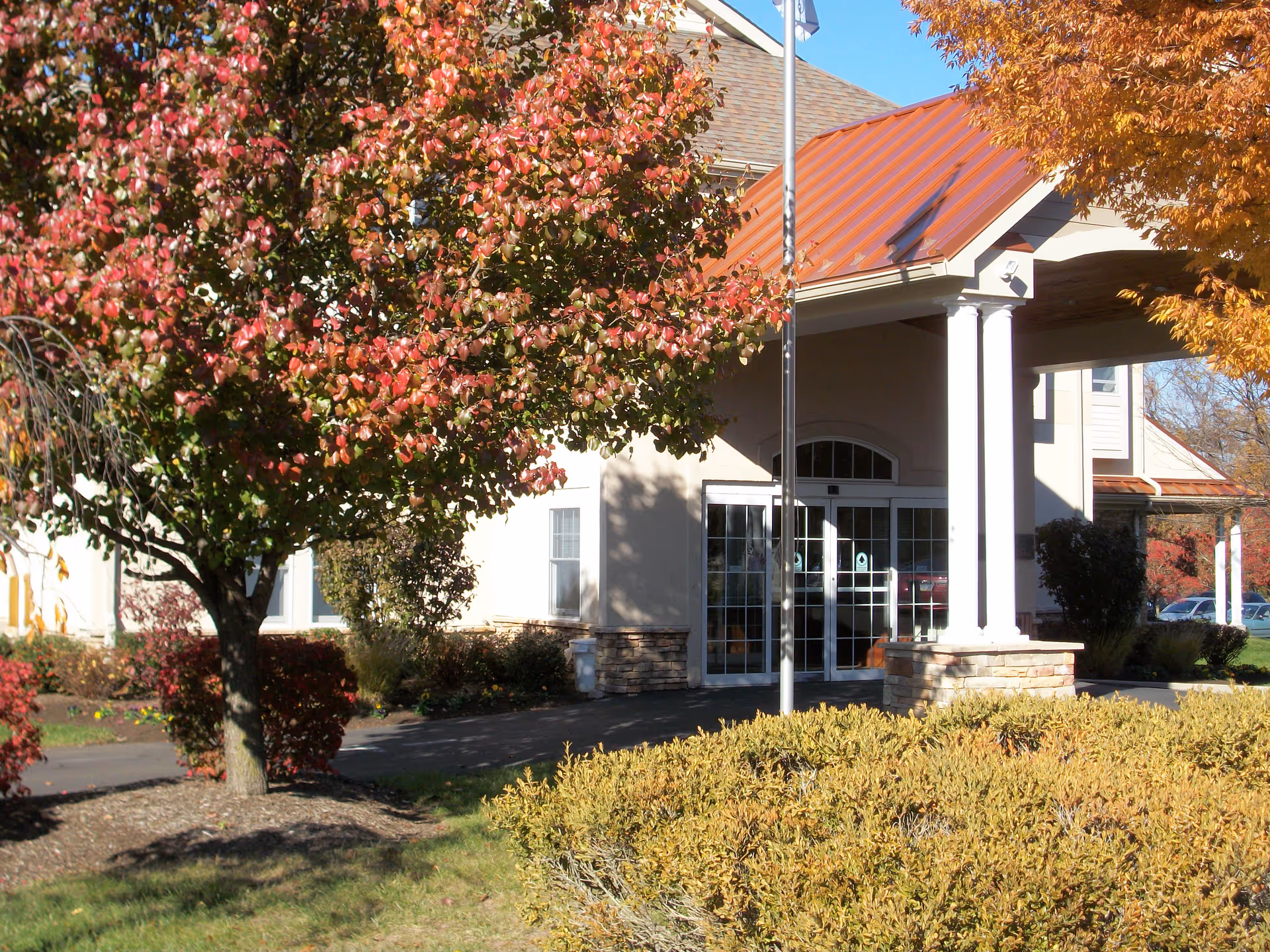 Entrance of Chandler Hall Health Services building with a covered porch supported by white columns, surrounded by trees with autumn foliage and landscaped bushes.