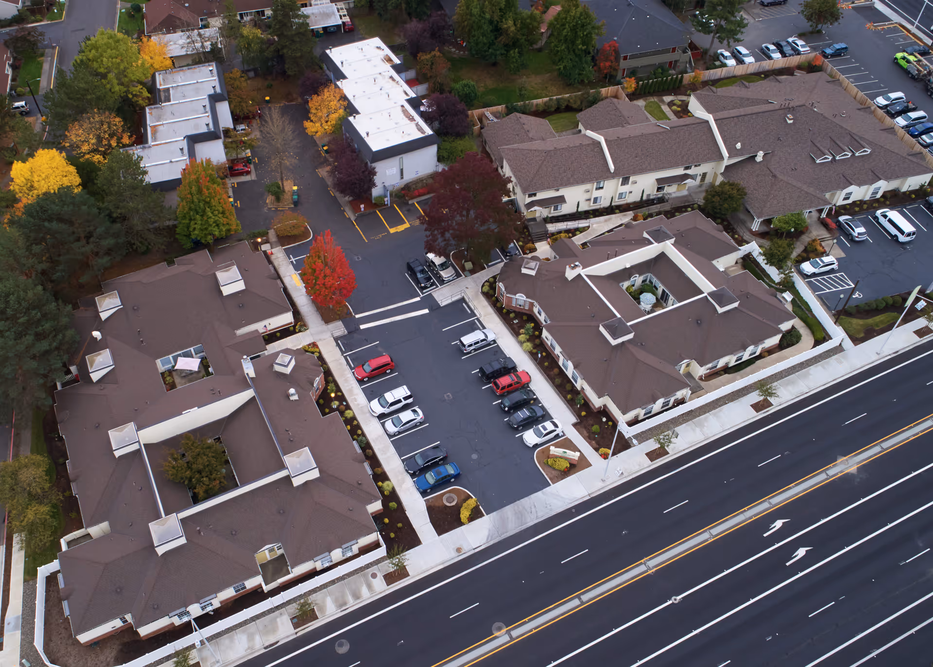 Aerial view of Farmington Square Beaverton senior living facility showing multiple buildings with brown roofs, parking lots with cars, surrounding trees with autumn foliage, and a multi-lane road in the foreground.