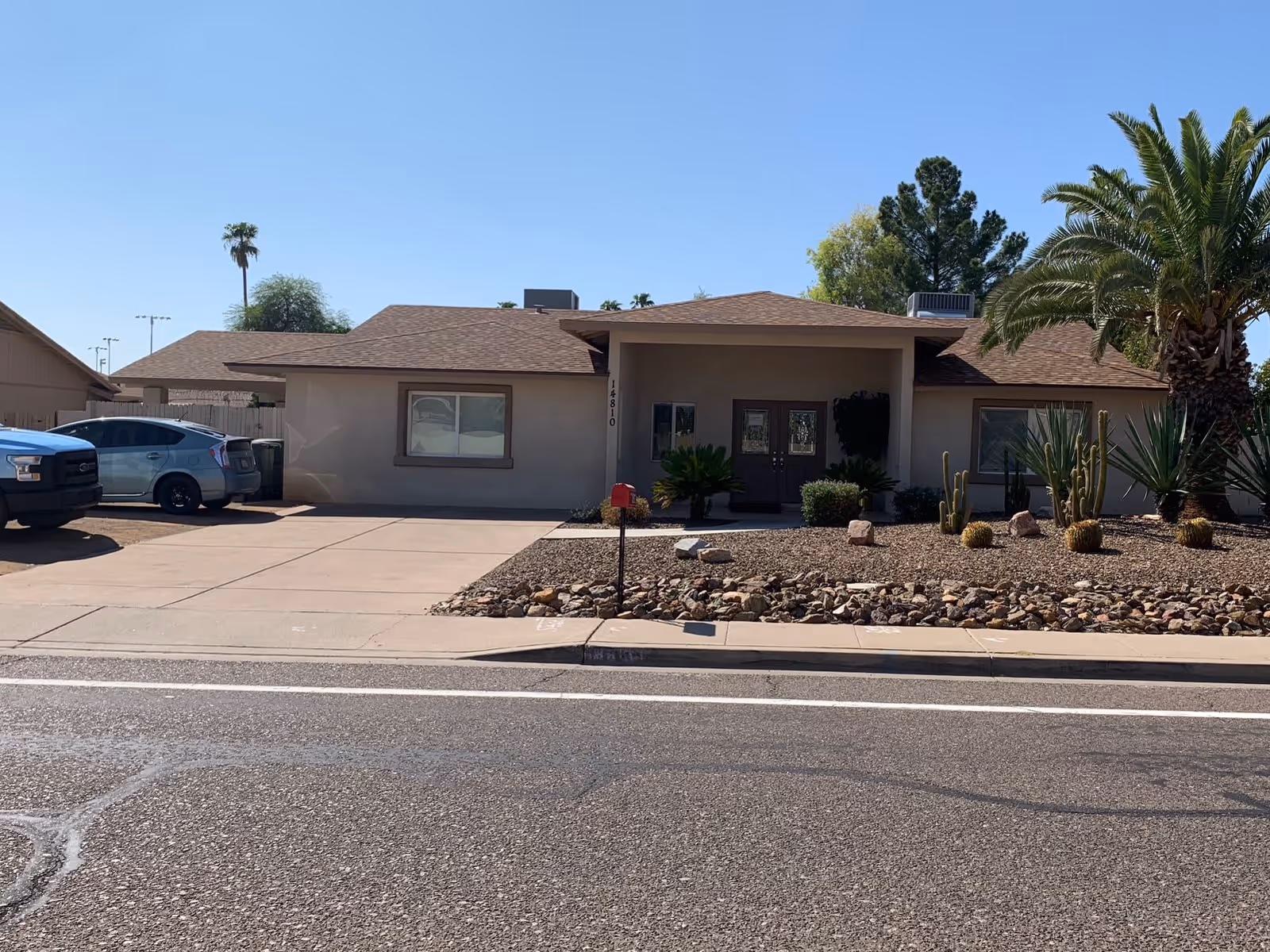 Front exterior view of a single-story residential building with a driveway and desert landscaping featuring rocks, cacti, and palm trees under a clear blue sky.
