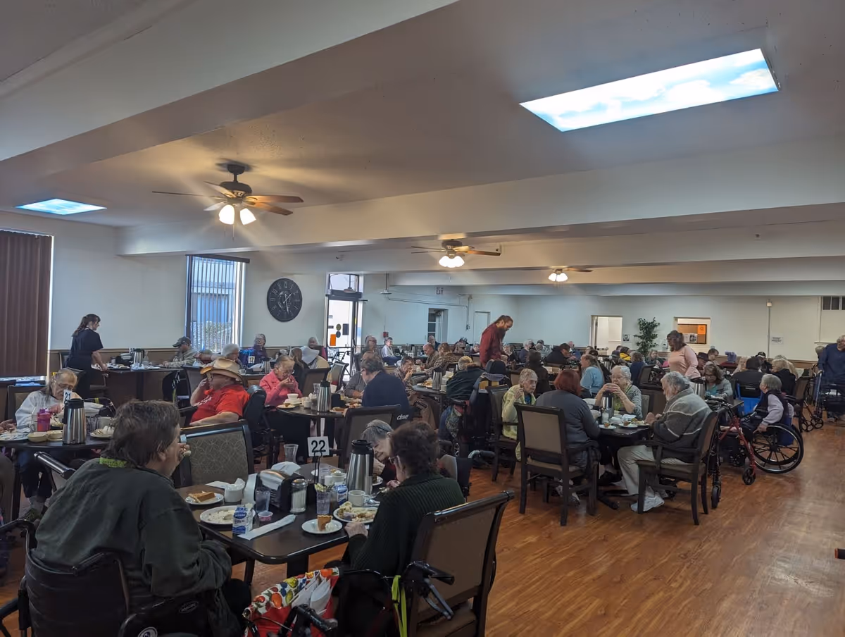 A large dining room filled with elderly residents sitting at tables eating meals. Several staff members are assisting and serving food. The room has ceiling fans, large windows, and a wooden floor.