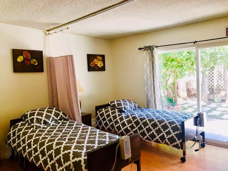 A bright room with two single beds featuring matching black and white patterned bedding. A privacy curtain is partially drawn between the beds. The room has light-colored walls with two floral paintings and a sliding glass door that opens to a garden area outside. A bedside lamp is visible between the beds.