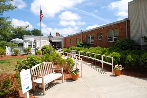 Outdoor view of a senior living facility entrance with a brick building, a concrete walkway with a white handrail ramp, a wooden bench, potted flowers, an American flag on a flagpole, and green shrubs under a partly cloudy sky.