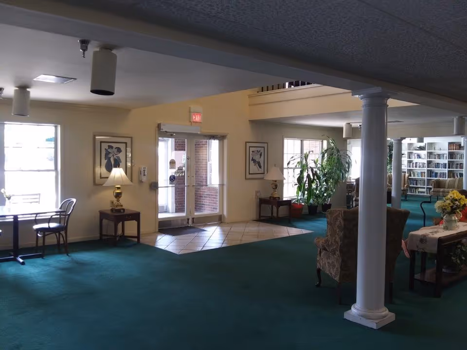 Spacious senior living lobby with green carpet, seating areas, plants, bookshelves, and double glass entrance doors.