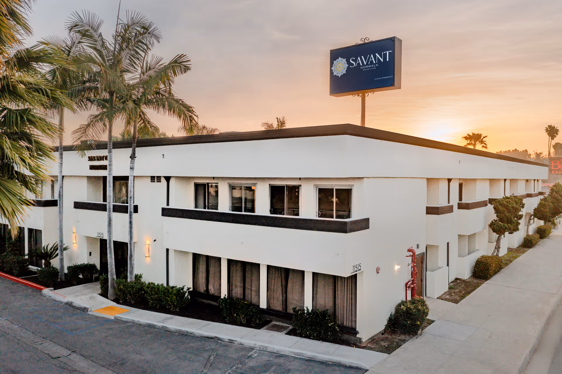 Two-story white Savant of Norwalk building with palm trees and a rooftop sign at sunset.