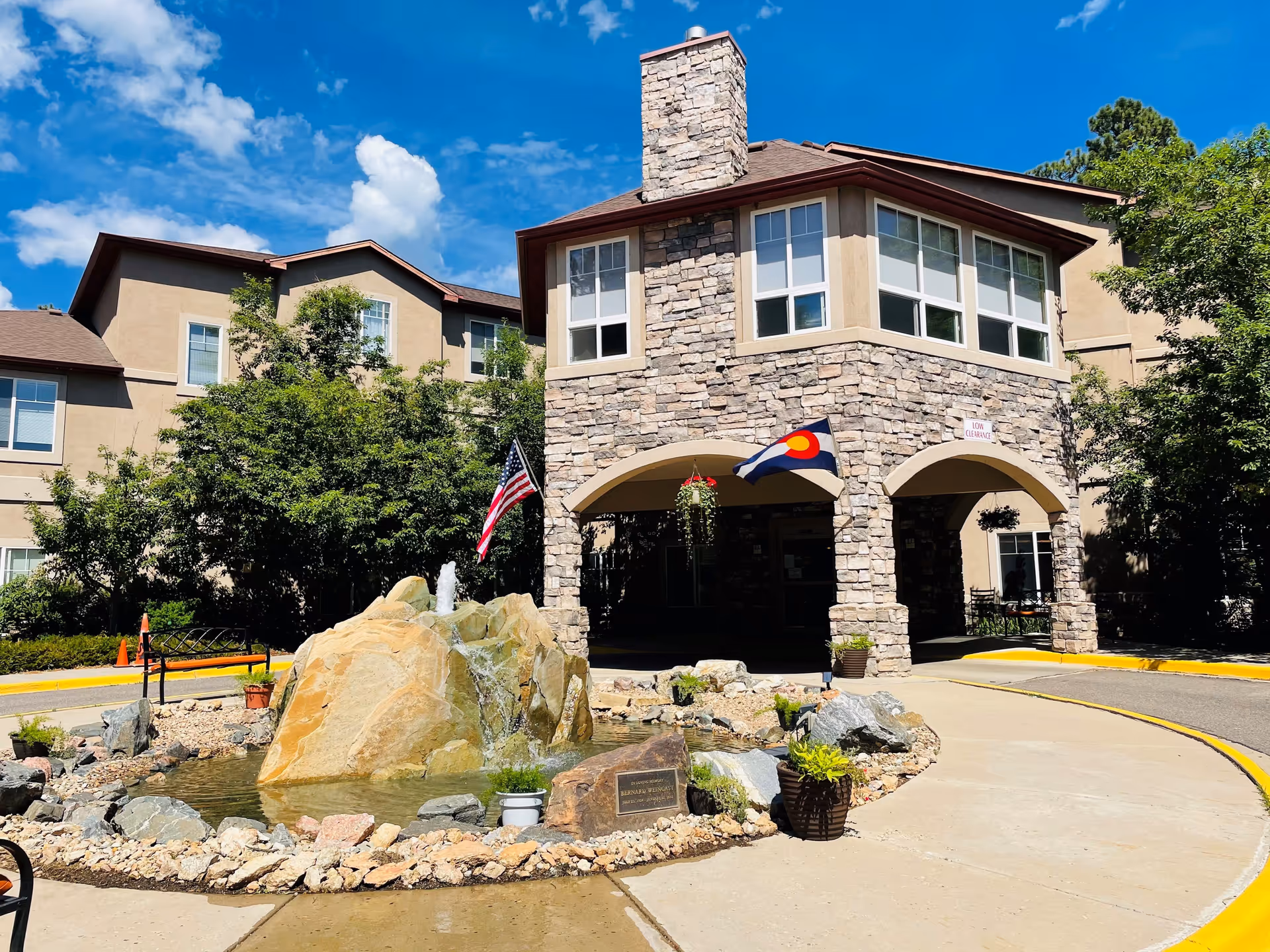 Stone-faced front entrance of Elk Run Assisted Living with a fountain, American and Colorado flags, and landscaping under a blue sky.
