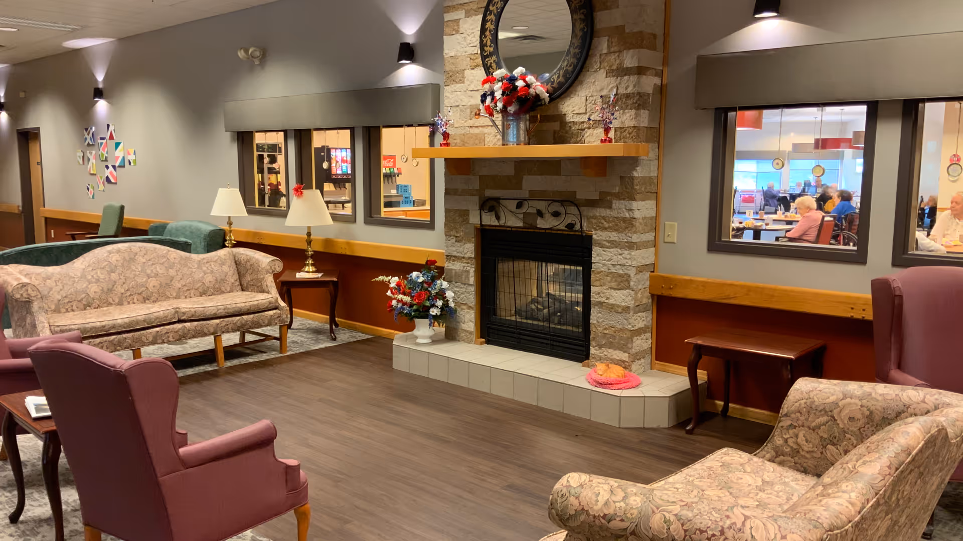 Cozy senior living lounge with upholstered sofas and chairs arranged around a stone fireplace and interior windows looking into a dining area.