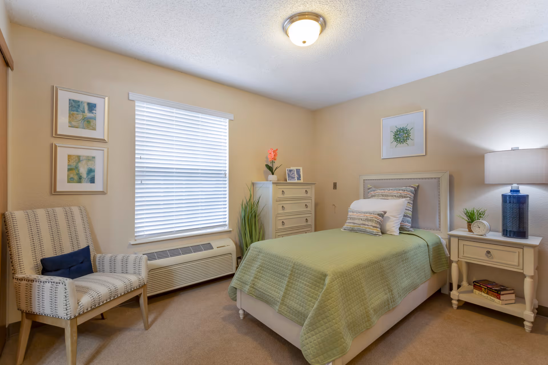 A cozy bedroom with a single bed covered in a green quilt and multiple pillows. Next to the bed is a white nightstand with a blue lamp, a small plant, and an alarm clock. There is a white chest of drawers with a pink flower and a framed photo on top. A patterned armchair with a dark blue cushion is placed near a window with white blinds. The walls are beige and decorated with framed artwork.