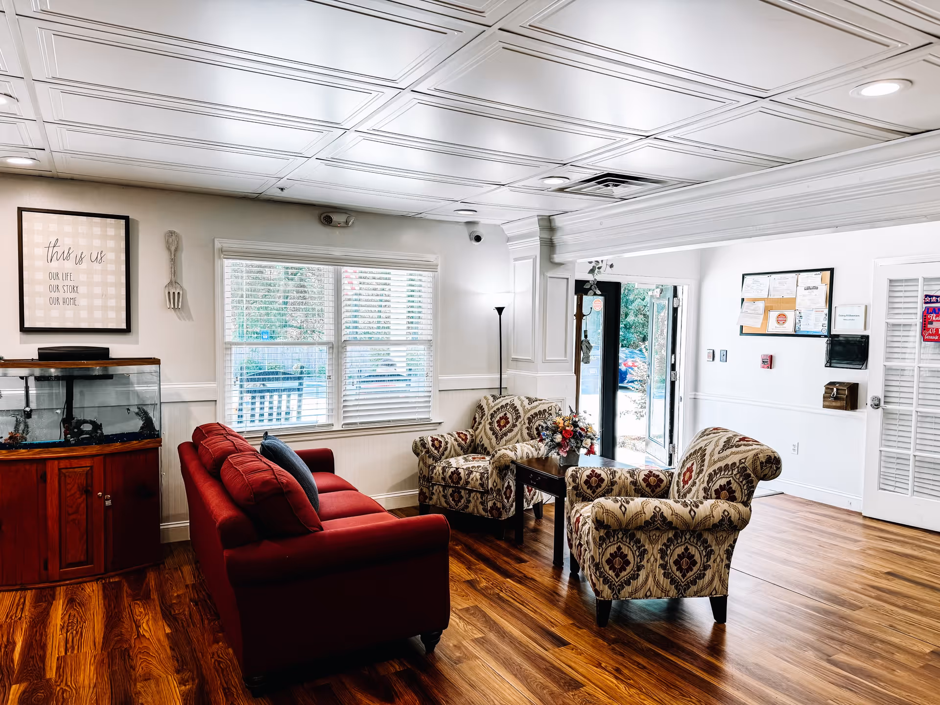 A bright sitting room with a red sofa, two patterned armchairs, a coffee table, fish tank and large window.