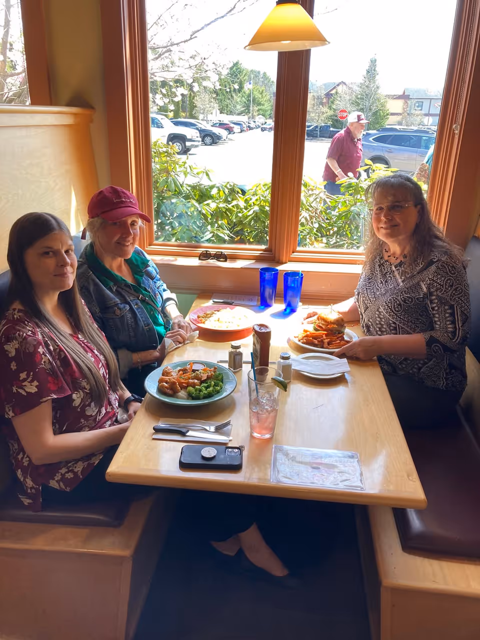 Three women sitting at a wooden booth table in a restaurant, enjoying a meal together. The table has plates of food, drinks, and condiments. Behind them is a large window showing a parking lot with cars and a man walking outside. A hanging light fixture is above the table.