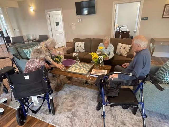 Four elderly individuals sitting and standing around a wooden coffee table in a cozy living room setting. Two are seated in wheelchairs, and two are seated on a sofa and chair. The coffee table has a chessboard, flowers in vases, and a book. The room has light-colored walls, a mounted TV, and doors leading to other rooms.