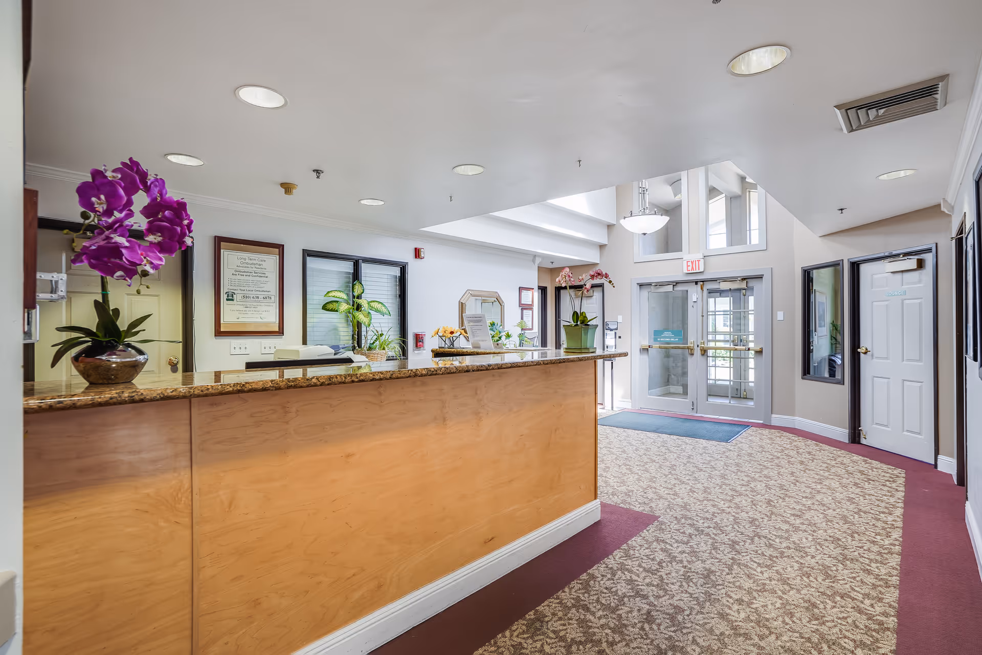 Reception desk and lobby area of a senior living facility with orchids, potted plants, and glass entry doors.